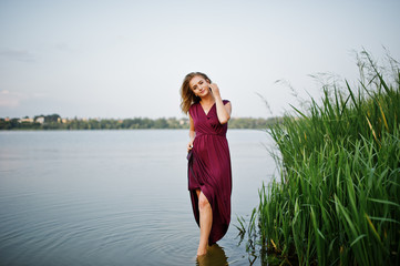 Fototapeta premium Blonde sensual woman in red marsala dress standing in water of lake with reeds.