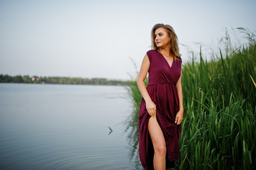 Fototapeta premium Blonde sensual woman in red marsala dress standing in water of lake with reeds.