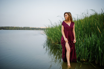 Fototapeta premium Blonde sensual woman in red marsala dress standing in water of lake with reeds.