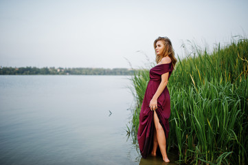Fototapeta premium Blonde sensual woman in red marsala dress standing in water of lake with reeds.