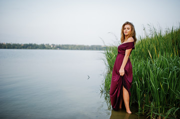 Fototapeta premium Blonde sensual woman in red marsala dress standing in water of lake with reeds.
