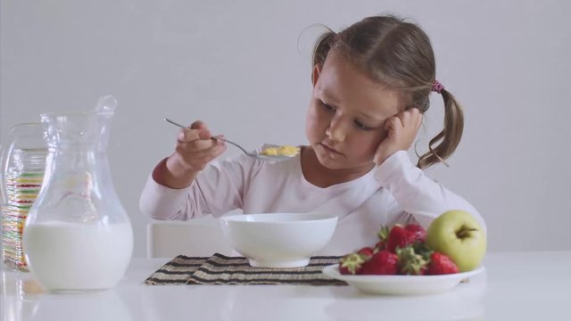 Portrait of sad small girl doesn't want to eat cornflakes with milk for breakfast. She is mixing her food with spoon sitting at table on white background.