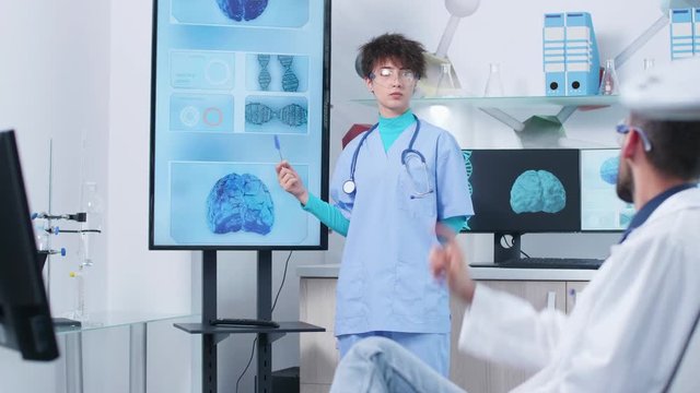 Young nurse with senior doctor in modern research facility analyzing 3D brain simulation on big screen TV and computer monitors