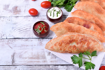 Golden Chebureki on a white plate. Sauces, vegetables and greenery around