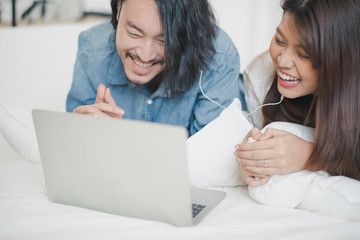 Young Asian man and woman are looking joke movie in laptop computer on the bed at home. the couple man and woman lie on the tummy and smiling with laugh when looking clip vdo. life at home concept.