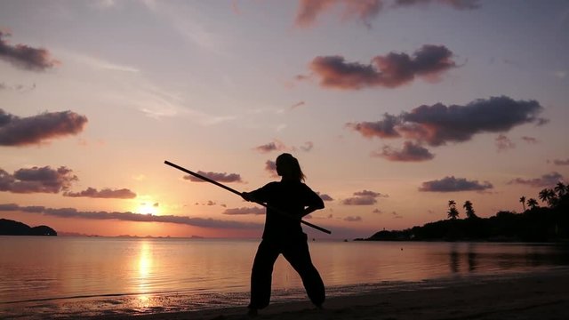 Silhouette of a young powerful woman standing on the beach with a kung-fu stick on the background of the sea at the sunset time.