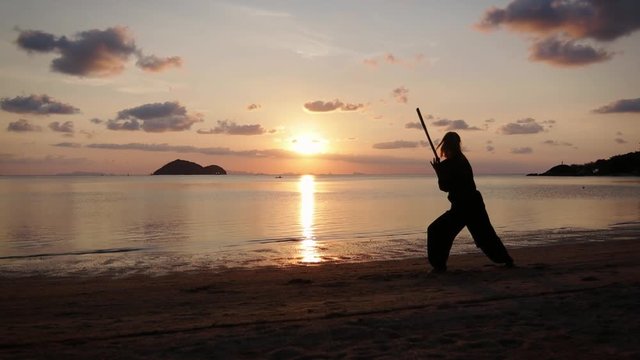 Silhouette of a young powerful woman on the beach doing a kung-fu exercises with a stick on the background of the sea.
