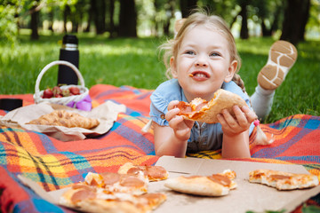 Little baby girl eating pizza and laughing outdoors on the background of green grass, summer...