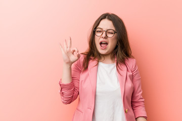 Young plus size business caucasian woman winks an eye and holds an okay gesture with hand.