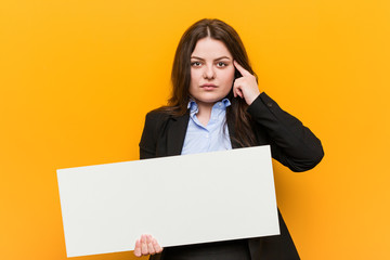 Young plus size curvy woman holding a placard pointing his temple with finger, thinking, focused on a task.