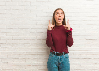 Young cool woman over a bricks wall surprised pointing up to show something