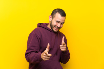 Colombian man with sweatshirt over yellow wall pointing to the front and smiling