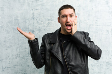 Young rocker man holds copy space on a palm, keep hand over cheek. Amazed and delighted.