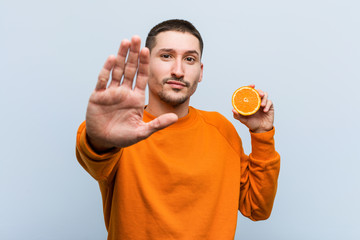 Young caucasian man holding an orange standing with outstretched hand showing stop sign, preventing...