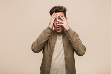 Young caucasian man wearing a brown jacket blink through fingers frightened and nervous.