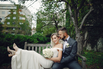 Fototapeta premium Beautiful bride in a long white dress. Handsome groom in a black suit. Couple in a summer park sitting on a bench