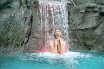 young sexy blond woman in pink bikini enjoys the falling water of the waterfall in the spa