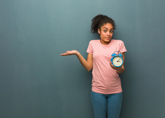 Young black woman doubting and shrugging shoulders. She is holding an alarm clock.