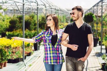 Beautiful young couple in casual clothes is choosing plants and smiling while standing in the greenhouse