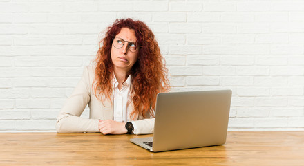 Young redhead curly woman working with her laptop confused, feels doubtful and unsure. - obrazy, fototapety, plakaty