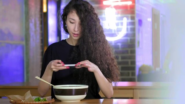 A lovely young Asian girl takes pictures on the smartphone pho soup at an Asian cafe. Chinese, Vietnamese or Japanese cafe or restaurant. Technology and social networks concept.