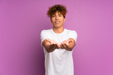 Young african american man over isolated purple wall holding copyspace imaginary on the palm to...