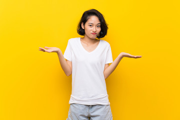 Asian young woman over isolated yellow wall having doubts while raising hands