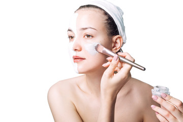 A woman on white background apply a grey clay face mask with a brush