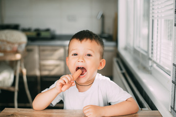 Portrait of a adorable little boy in a white t-shirt at the table eating a sausage with his hands