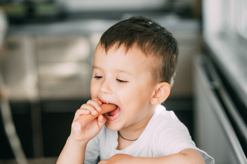 Portrait of a adorable little boy in a white t-shirt at the table eating a sausage with his hands