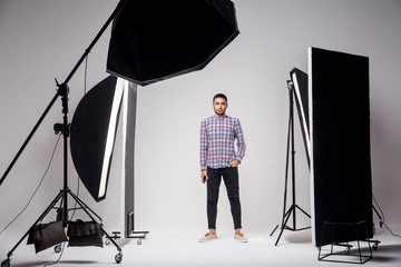 Professional photography studio showing behind the scenes lights. fashion handsome young man model at studio in the light flashes, standing and looking at camera. indoor studio shot on grey background