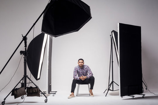 Professional photography studio showing behind the scenes lights. fashion handsome young man model at studio in the light flashes, sitting and looking at camera. indoor studio shot on grey background