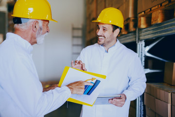 Two male Caucasian employees in white uniforms and with yellow helmets on heads talking about sale growth while standing in warehouse. Younger one holding tablet while older one pointing at chart.