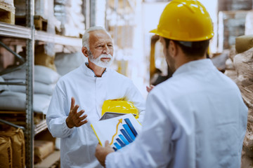 Two employees talking about shipping inn warehouse. Older one holding tablet while younger one holding chart.