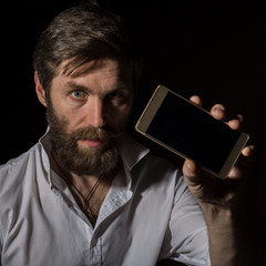 handsome bearded man shows his phone with fascinating smile on a dark background.