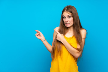 Fototapeta premium Young woman with long hair over isolated blue wall frightened and pointing to the side