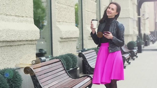 young attractive girl in black leather jacket and pink skirt. stands near the benches, hand holding coffee in a paper Cup and smiling. city street in the background