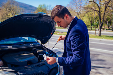 A handsome businessman wearing blue blazer lifting up the hood of his car and checking the oil level on a sunny day parked on a busy city boulevard.