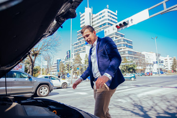 Obraz premium A handsome businessman wearing blue blazer lifting up the hood of his car and checking the oil level on a sunny day parked on a busy city boulevard.