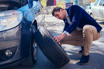 A tired businessman on a sunny day is taking a tire out of his car in order to be able to change his flat tire.
