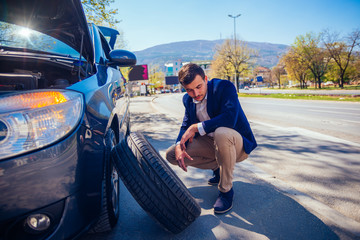 Angry businessman trying to change his flat tire with a socket wrench while crouching down next to his car.