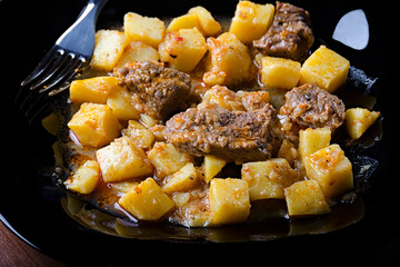 braised potatoes with meat and gravy on a black plate, closeup 45 view