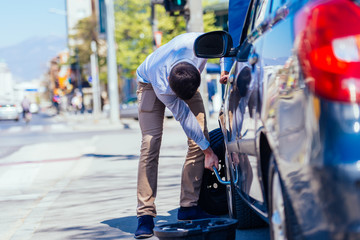 A sweaty tired young entrepreneur is wiping the sweat off his forehead while trying to change a flat tire on a beautiful sunny day