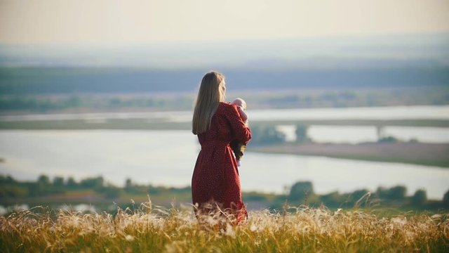 A blonde woman playing with her little baby on the field on a background of the river
