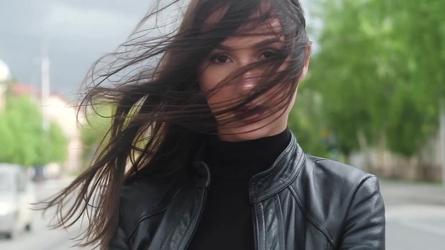 attractive sexy woman with long dark hair and black jacket walks down  street of the city on Sunny summer day. looks intently at camera, turns his head to side. strong cold wind ruffled her hairstyle.