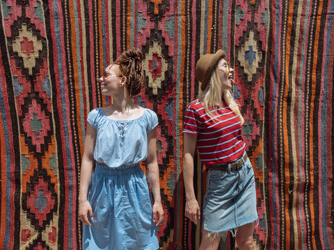 Two tourist girl friends posing on street in front of old east oriental carpets.