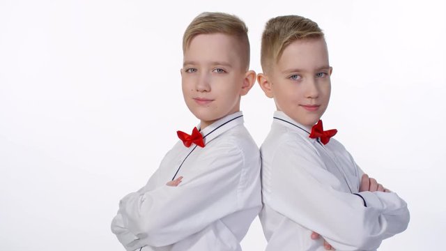 Waist-up shot of 10-year-old Caucasian identical twins, dressed up in white shirts and red bowties, standing back to back against white background in studio and posing for photos with crossed arms