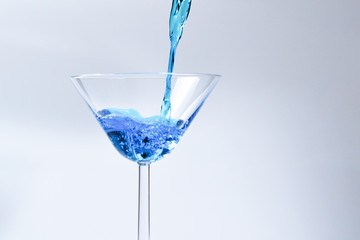 Cocktail with blue liquid in glass. Glass with blue water pouring with liquid with splashes and drops. Martini glass filling with alcohol with splashes on white background. Refreshing drink concept.