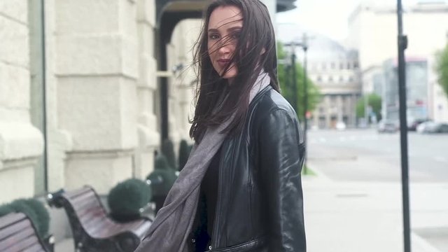 beautiful cheerful young girl with long dark hair, grey scarf, black leather jacket and jeans. Looks in the frame, smiles, turns away and goes on a city street. Long hair fluttering in the wind