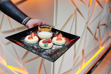 Man holds black square plate with homemade cheese pancakes with fresh berries, honey and sour cream on a beautiful light background with geometric lines. Close-up. Space